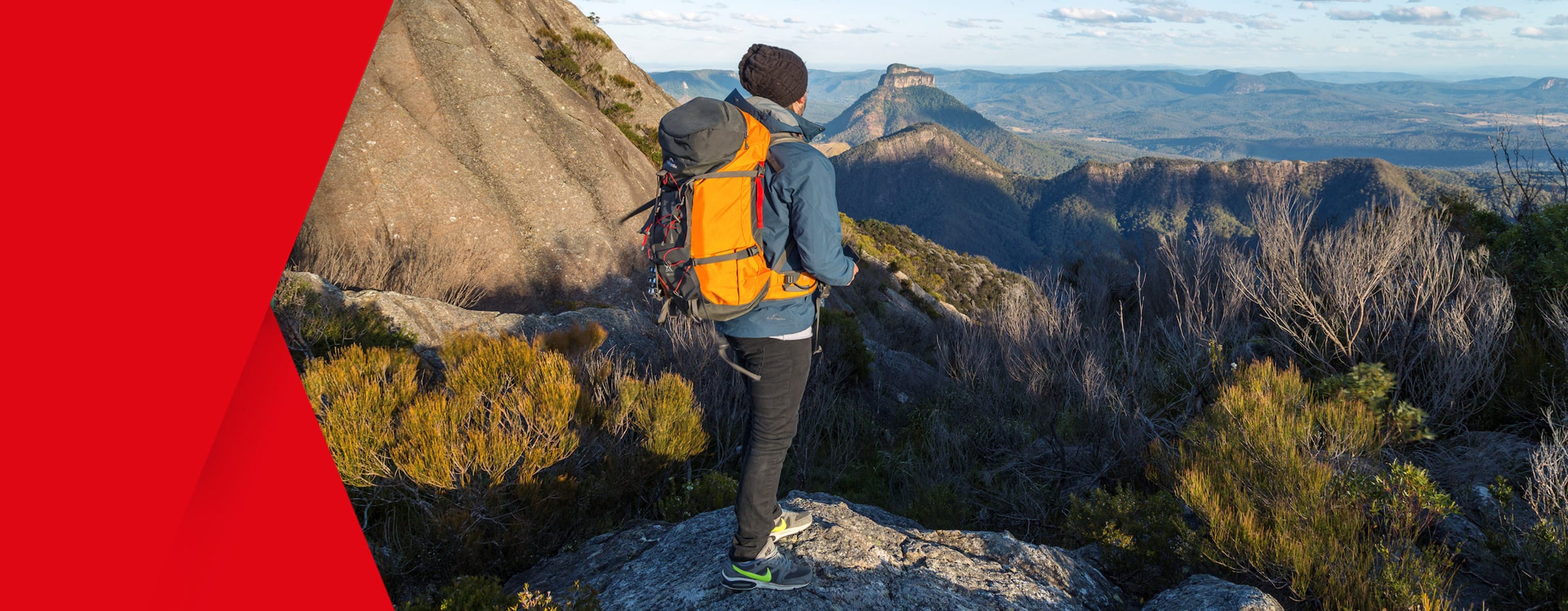 A man in warm hiking gear and backpack stands on a rock overlooking a bushy mountainous range far in to the distance