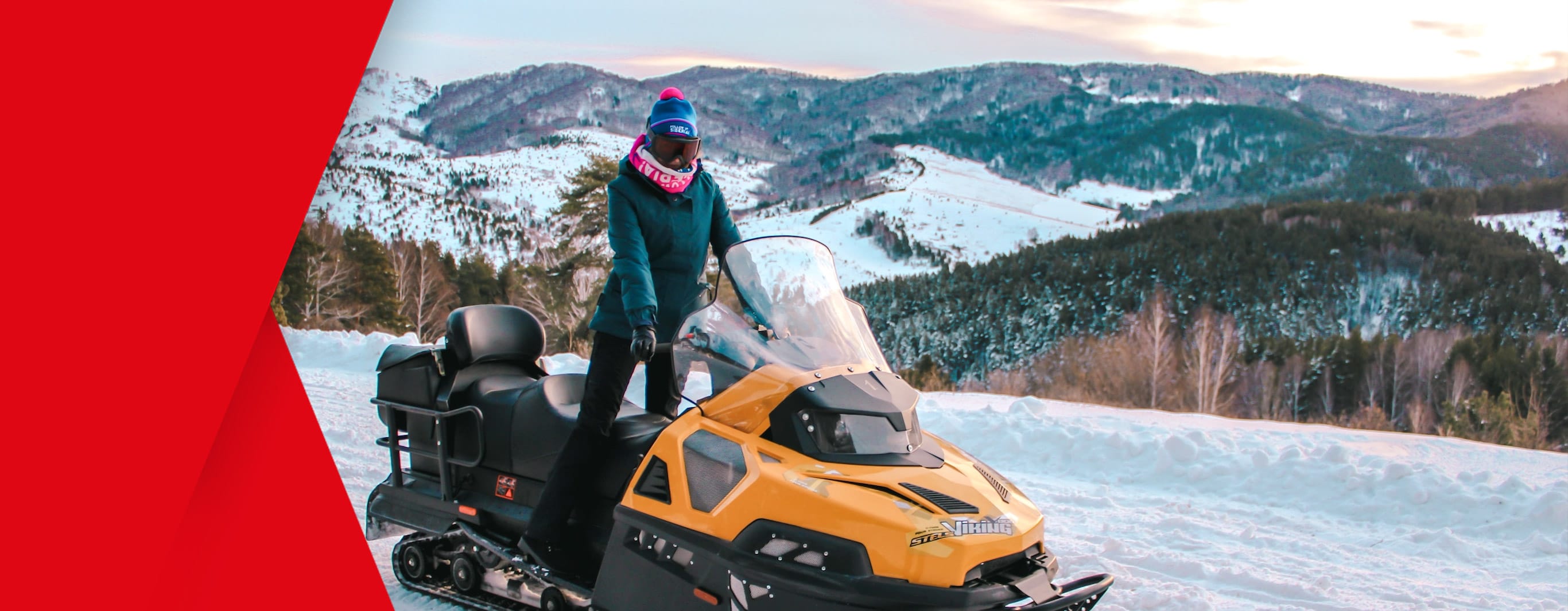 A woman wearing thick skiing clothing and full-face glasses poses upright upon a stationary snowmobile in the midst of the frozen wilderness