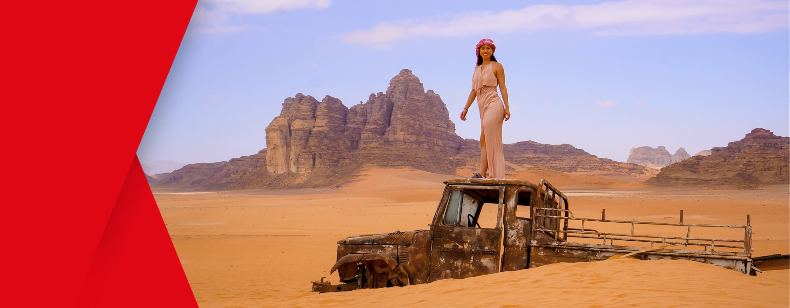 A woman in a billowing full length dress and wearing a floral crown smiles for the camera as she stands atop an old, rusty, abondoned pickup truck that has been semi-consumed by the desert sands. In the distance we see rocky outshoots rising from the sand to form a dramatic cliff backdrop