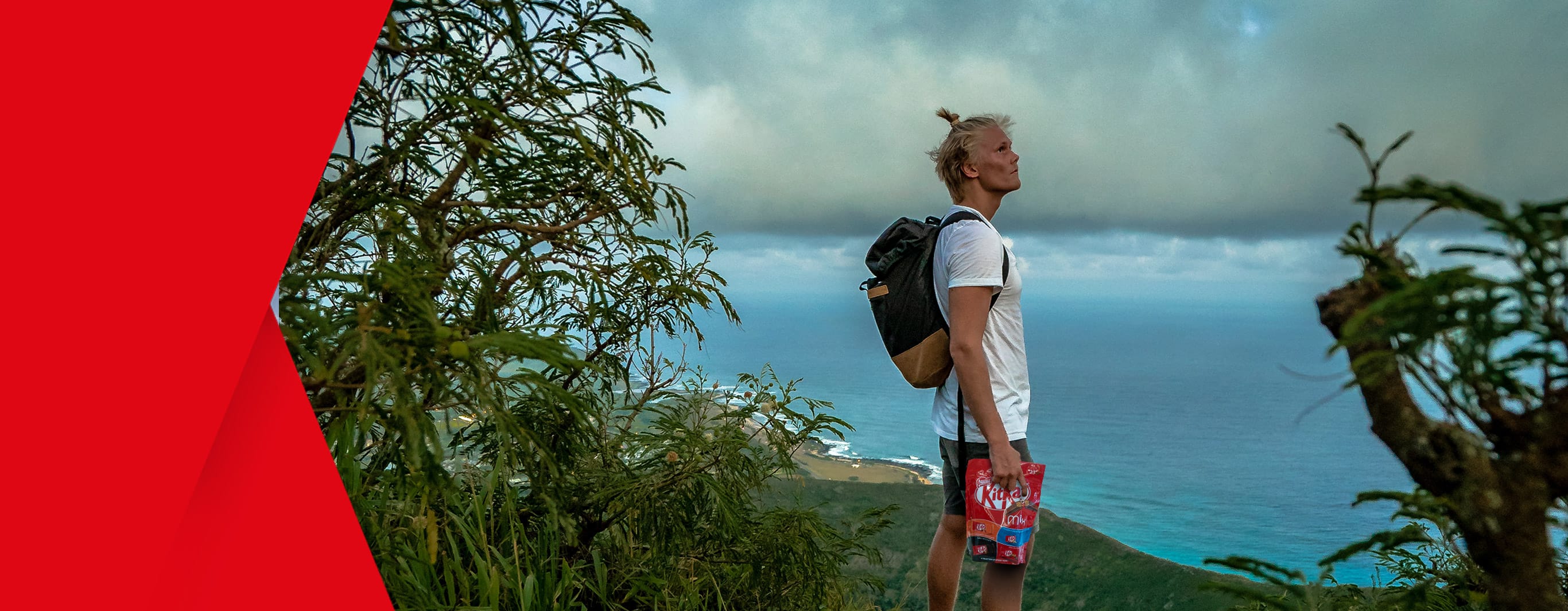 A man holding a bag of KitKat chocolates and surrounded by bushes stands at the edge of high vantage point, overlooking a wide vista of the sea and tropical shoreline in the below