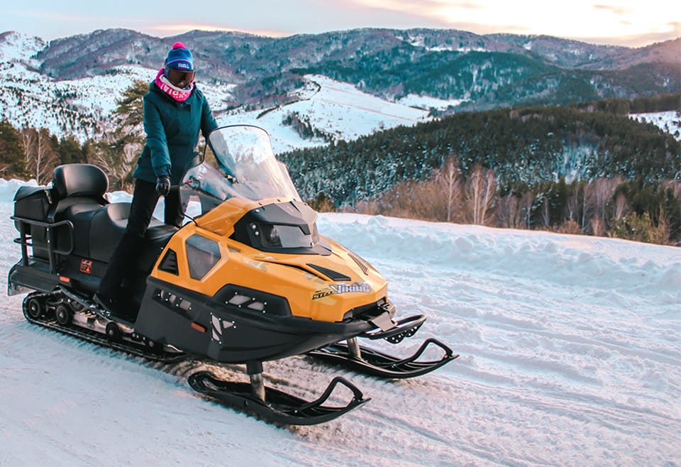 A woman wearing thick skiing clothing and full-face glasses poses upright upon a stationary snowmobile in the midst of the frozen wilderness