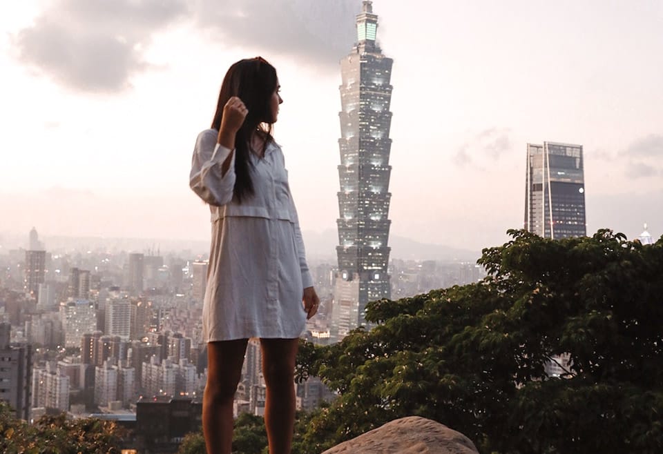 A woman in the foreground poses in front of the Taipei 101 skyscraper, which looms over the city of Taipei, Taiwan