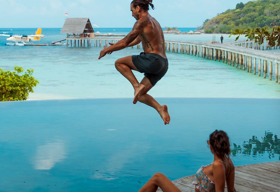An athletic man leaps in to a deep blue swimming pool while a female leans back and watches. In the near background is light blue ocean with wooden walkway leading to a seaplane