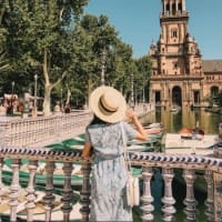 A woman in the foreground holds her rimmed hat as she stands on a bridge overlooking the narrow canal of the Plaza de España. In the background is part of the balustraded balconies of a Renaissance Neo-Moorish-style building, with gondolas visible in the canal below