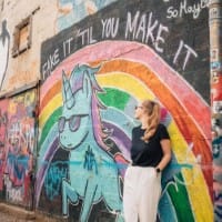 Woman leaning against a wall with a colourful painted murial that reads 'Fake it til you make it'