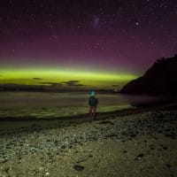 A lone figure stands on a beach at night watching in the distance the spectacular sight of Aurora Australis
