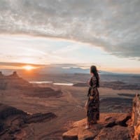 A woman in a long floral dress stands at the precipice of a cliff overlooking the spectacular and desolate canyon below, as the sun rises in the distance